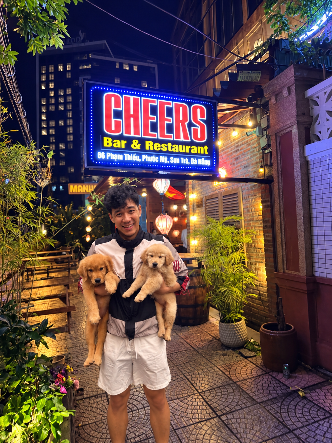 Danny holding two Golden Retriever puppies beneath the illuminated Cheers sign in Da Nang